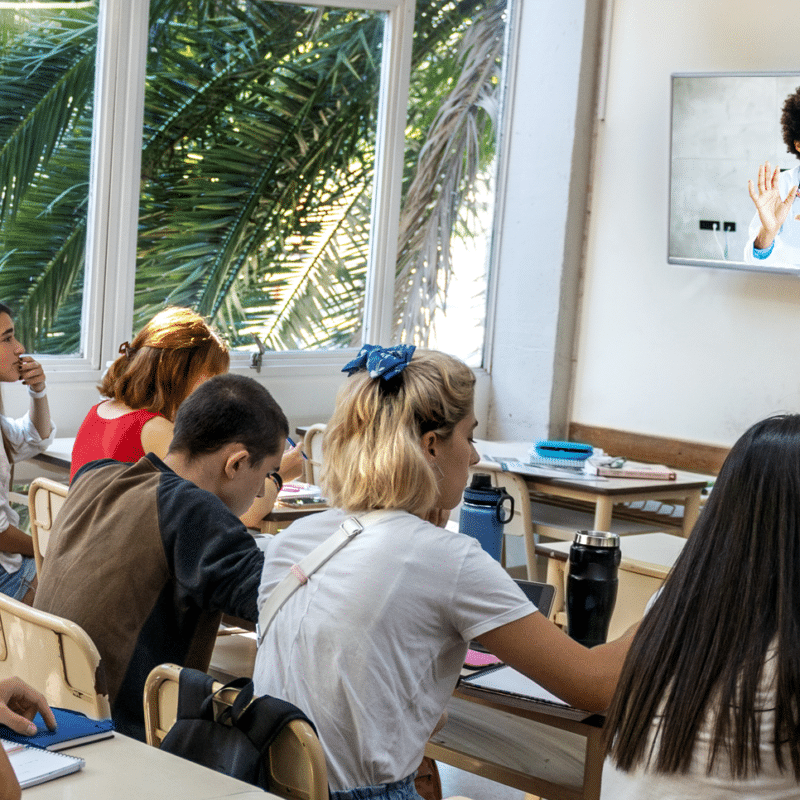 Photo of students in a classroom taking notes and watching a television on the wall that is displaying a Kids Making Sense scientist gesture and speak.